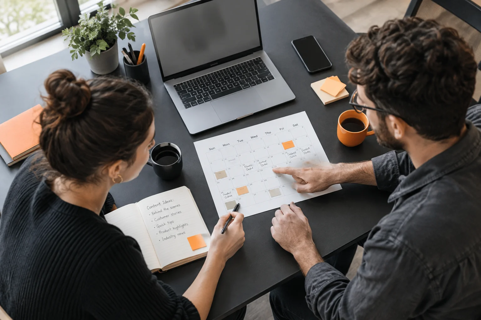 Overhead view of a small business owner and team member planning a social media content calendar at a charcoal desk with orange-accented stationery.