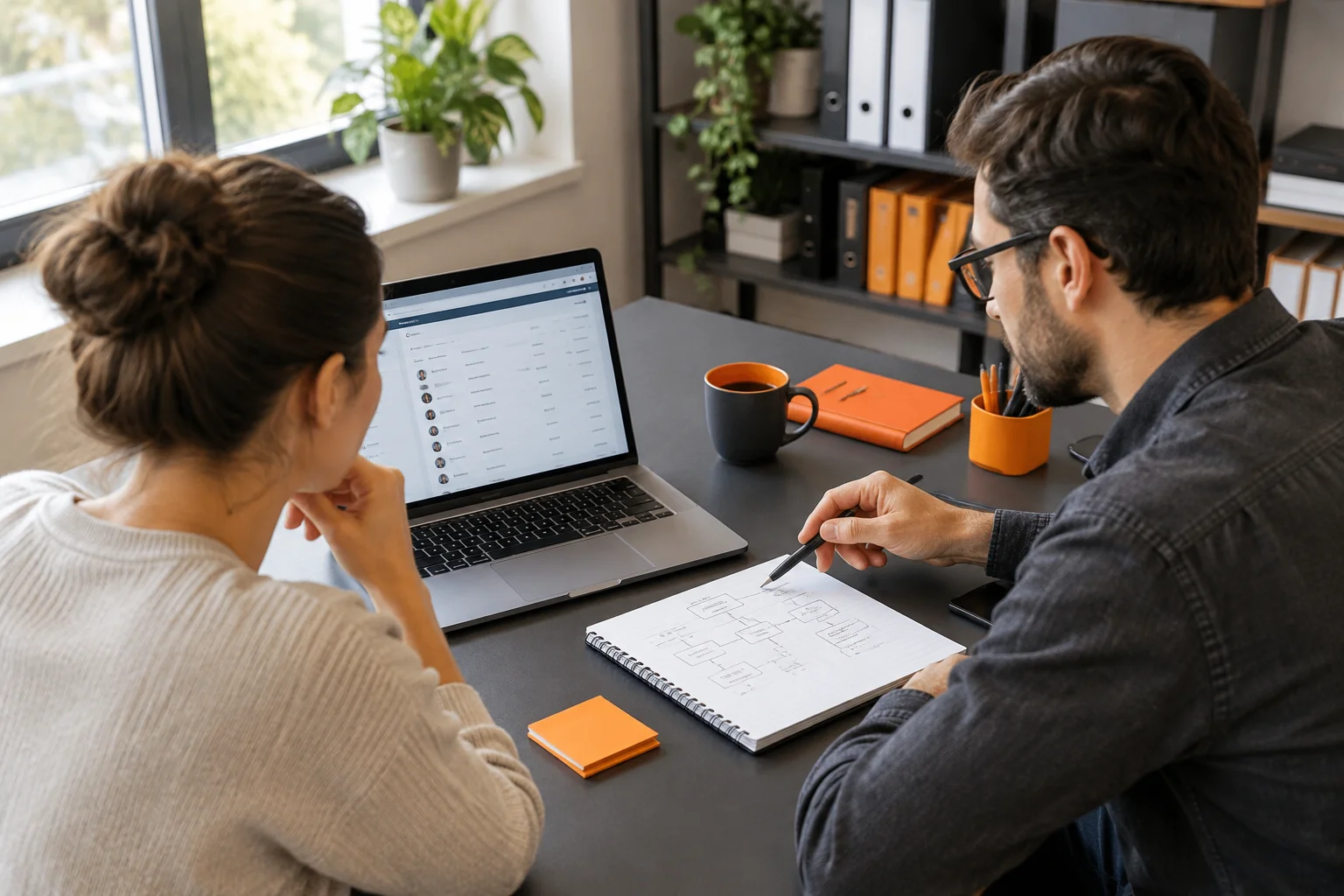 Small business owner and web developer reviewing CRM integration notes at a desk with a laptop, orange-accented stationery, and natural daylight.